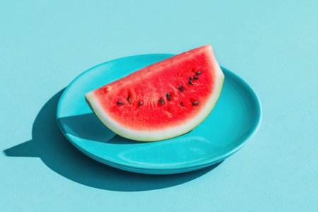 A vibrant red watermelon slice with visible seeds rests on a bright turquoise plate, casting sharp...