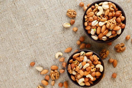 A mixture of cashew nuts, almond nuts, pistachios, hazelnuts and walnuts in a wooden cup against the background of burlap fabric. Nuts as structure and background, macro. Two cups of nuts.の写真素材