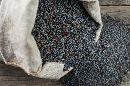 Poppy seeds in a burlap bag on a vintage wooden gray background. In country style. The tasty and useful seeds rich with protein and oils. It is poured out of a bag. Calcium content leaderの写真素材