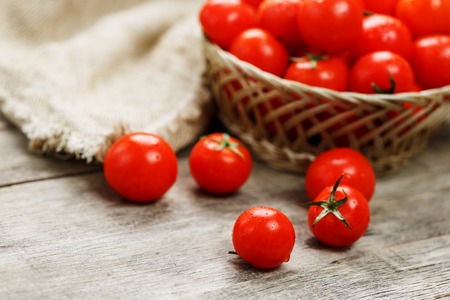 Fresh red tomatoes in a wicker basket on an old wooden table. Ripe and juicy cherry tomatoes with drops of moisture, gray wooden table, around a cloth of burlap. In a rustic style.の写真素材