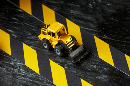 Yellow toy bulldozer on a black background texture. Black and yellow fence tape against the background of the CONCEPT. Business, repair, services.の写真素材