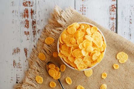 Bowl with golden flakes on a linen, on a light wooden table, next to a spoon. Rustic country style. View from above. White cup, morning healthy breakfast of cornの写真素材