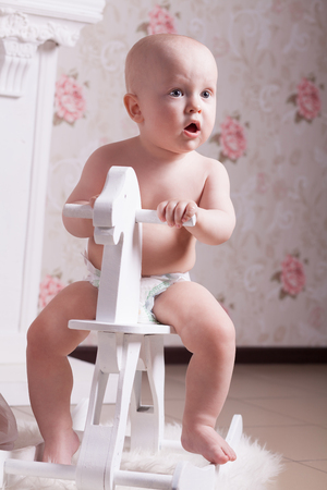 Funny baby in diapers is sitting on a white, wooden, toy horse and looks in surprise somewhereの写真素材