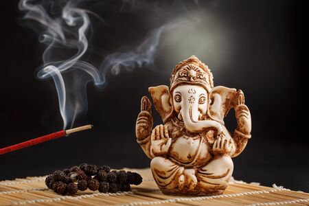 Hindu god Ganesh on a black background. Rudraksha statue and rosary on a wooden table with a red incense stick and incense smoke. Copy spaceの写真素材