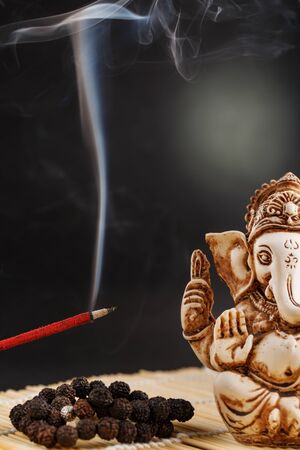 Hindu god Ganesh on a black background. Rudraksha statue and rosary on a wooden table with a red incense stick and incense smoke. Copy spaceの写真素材