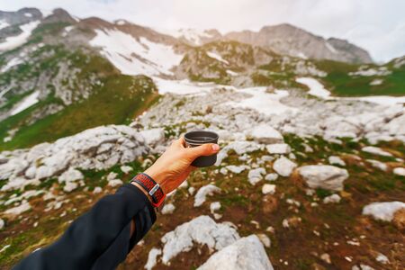 A hand with a mug of clear water against the backdrop of a snow-covered mountain landscape. The traveler drinks.の写真素材