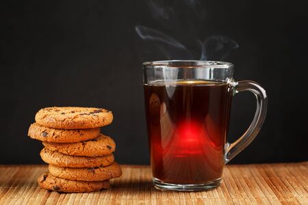 A pile of oatmeal cookies with chocolate chips and a mug of fragrant black hot tea in on a bamboo substrate, on a dark background. Handmade cookies for a healthy breakfast. Close-upの写真素材