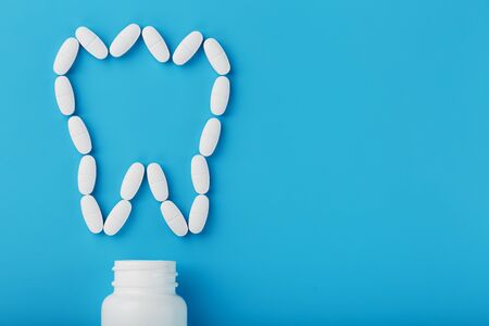 Tooth made of white vitamins with calcium on a blue background. Vitamins spilled out of a tooth-shaped jar. Useful vitamins for bones, teeth and hair. Ca, D3.の写真素材