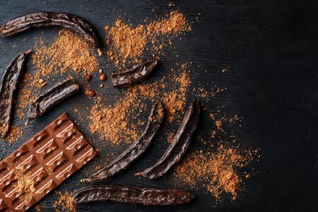 The pods of the carob chocolate bar on a black background. Sweets, chocolate and confectionery made of carob fruit. Replace cocoa and sugar with useful . Top view, free space for textの写真素材