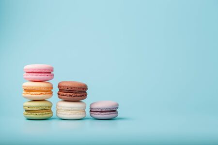 Macaroni cookies of different colors are arranged in the shape of pyramid steps on a blue background. Free space. The concept of minimalismの写真素材