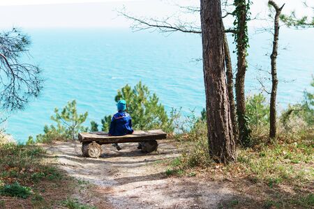 A lonely child is resting on a bench in the forest with a view of the sea. The concept of loneliness, freedom and isolation.の写真素材