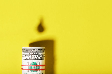 A roll of American bills with the shadow of an oil barrel and a drop of oil on a yellow background....
