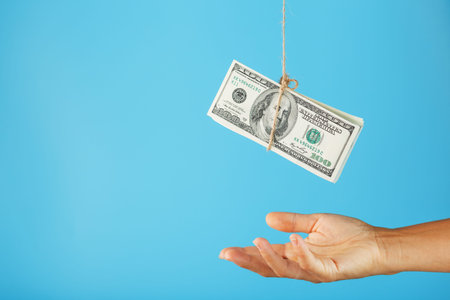 A wad of dollars on a string above a human palm on a blue background.