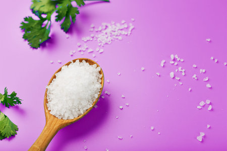 Large white sea salt in a wooden spoon on a pink background, with salt and herbs scattered around...