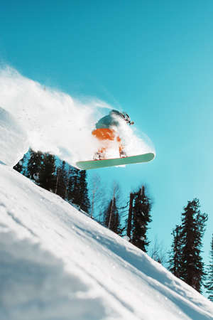 A snowboarder jumps from a high snow-covered springboard in a forest against a blue sky. Extreme sportsの写真素材