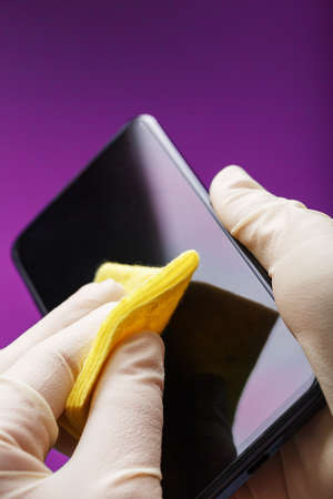 Cleaning a smartphone with a yellow napkin from dirt and viruses in rubber gloves on a pink background. Cleaning a mobile phone from viruses, isolated.の写真素材