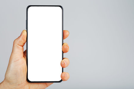 Hand holds black smartphone with blank white screen and modern frameless design isolated on gray...