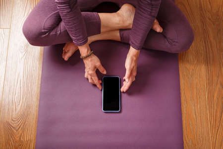 A girl practicing yoga and meditation at home on a lilac carpet using a smartphone and a mobile app....