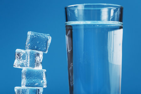 A glass of refreshing water and a tower of ice cubes on a blue background. A refreshing drink on hot...