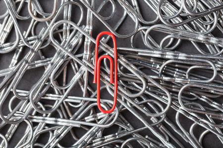 A red paper clip stands out against a textured background of silver paper clips. Concept