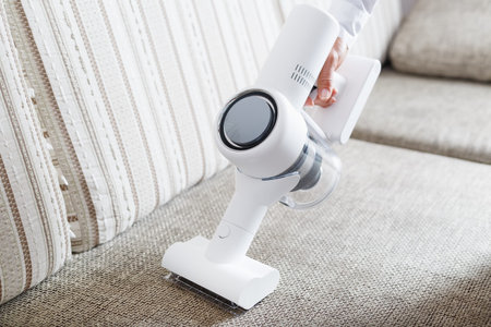 A man's hand holds a modern wireless vacuum cleaner for cleaning the sofa in the house.