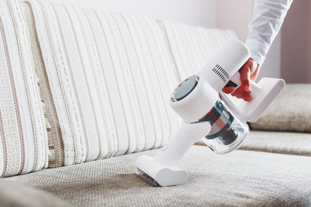 A person uses a white cordless vacuum cleaner to clean the sofa in the living room.