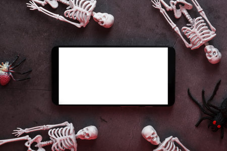 Decorative skeletons lie near the smartphone on a dark background. Halloween decoration, Top view,...