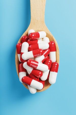 A handful of Red and white pill capsules in a wooden spoon on a blue background. Close-up of Free...