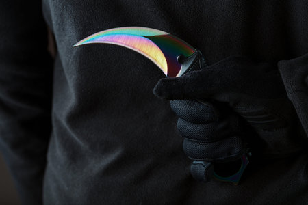 A criminal with a sharp knife hidden behind his back. Curved sharp blade of rainbow-colored kerambit