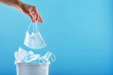 A hand throws a used protective mask into a trash can filled with masks from a virus on a blue...