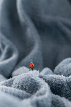 A figurine of a miniature mountain climber, set on a soft fleece surface that mimics a snow-covered landscape, with a hazy backdrop of a mountain or an icy wall. A conceptual miniature scene depicting a mountain climber on snow-covered mountain peaks.の写真素材