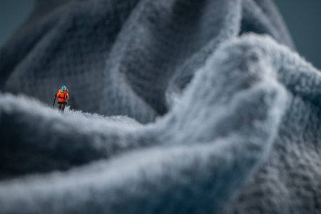 A miniature human figure in an orange jacket, blue hat, and ski poles stands on a fuzzy fleece surface resembling a snow-covered surface. In the background is a blurred image that could be a mountain or an ice wall. This conceptual miniature diorama depicts a climber on snow-capped mountain peaks.の写真素材