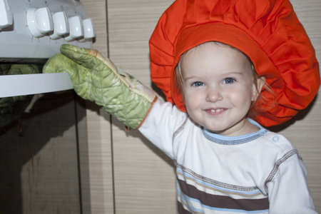 Baby with gloves and hat is opening the ovenの写真素材