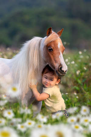 A child hugs a miniature horse outdoors. Little cute girl posing with a small horse in a field in spring.の写真素材