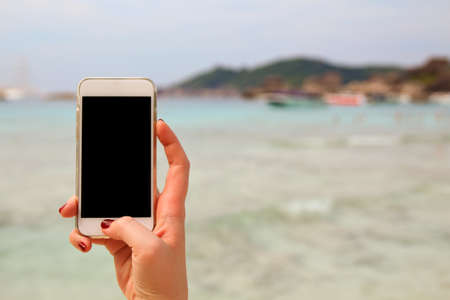 Female hand holding white mobile phone with blank screen on a tropical beach.の写真素材