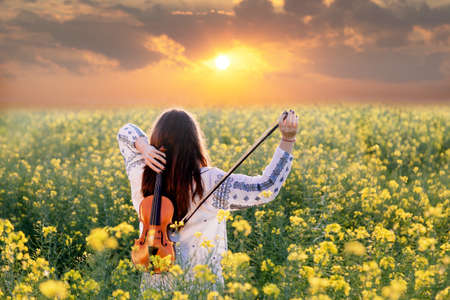 Young woman playing violin in a field at sunset. Young joyful girl wearing traditional romanian blouseの写真素材