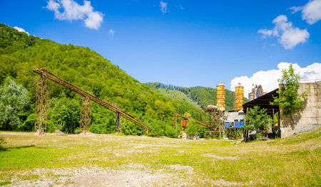 Old cement factory mine abandoned and forgoten in a desserted area with green woods and a blue cloudy sky on a sunny dayの写真素材