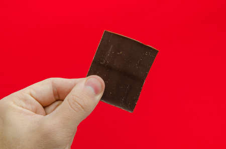Chocolate chunk being held by a hand over a red alerted dangerous background suggesting weight gain and problems due to eating sugar filled sweet dessert and convincing you to loose weight and go on a dietの写真素材