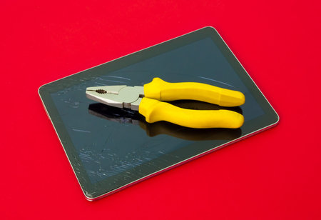 A pair of pliers on a broken touch screen tablet on a red alerted serious dangerous important background suggesting quick and expensive repair services for broken electronics in quick timeの写真素材