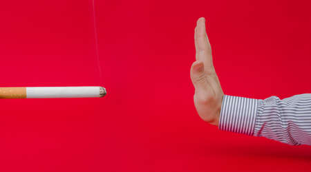Say no to smoking suggested by a business man refusing a giant lit smoking cigarette trying to loose weight and cut off an unhealthy habbit on a red dangerous serious alerted backgroundの写真素材