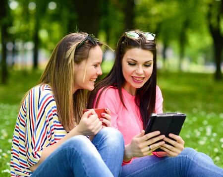 Young women drinking coffee and browsing internet on tablet at park and enjoying summer dayの写真素材