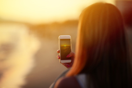 Tourist hand holding smart phone and taking photo of beautiful beach sunsetの写真素材