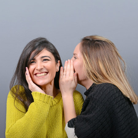 Two woman friends whispering secrets against gray background