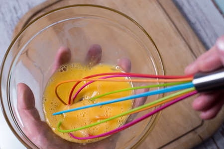 Egg yolk and white in glass bowl and wire whisk over dark weathered wooden table, cutting Boardの写真素材