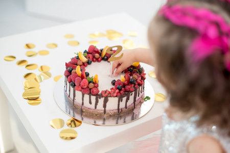 birthday cake with fruits and nuts on a light background with confetti, with the number 2 and the baby from the back.の写真素材