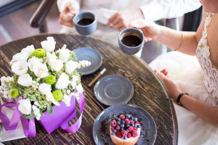 Hands of young couple eating cakes with coffee on wooden table with gift box and flowers.の写真素材