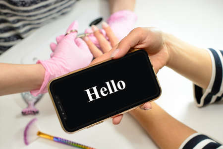 Woman using mobile phone, Hello on the screen, woman in salon receiving a manicure and nail care procedure by beautician. Female getting nails done by manicurist at beauty salon.の写真素材