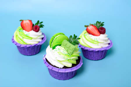 Three cupcakes with whipped cream, chocolate bar, strawberry ,decorated macaroons on blue background. Picture for a menu or a confectionery catalog. with space for textの写真素材
