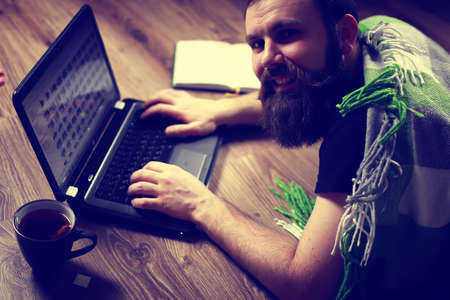 bearded handsome man lies on the floor wrapped in a plaid, and working at a laptopの写真素材