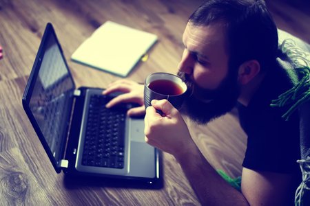 bearded handsome man lies on the floor wrapped in a plaid, and working at a laptop